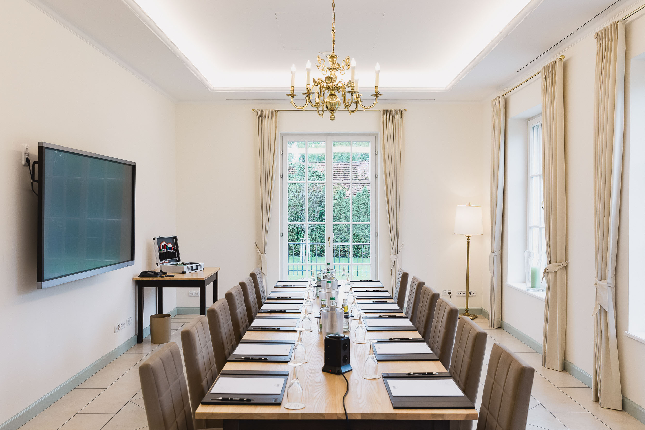 kleiner Tagungsraum mit bodentiefen Fenster, hellen Wänden. In der Mitte eine lange Tafel mit braunen Stühlen herum, einen Fernseher an der Wand. kleiner Tagungsraum mit bodentiefen Fenster, hellen Wänden. In der Mitte eine lange Tafel mit braunen Stühlen herum, einen Fernseher an der Wand.